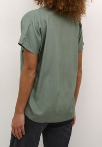 Mørkegrøn kortærmet top lavet af glat stof, med en rund halsudskæring og en afslappet pasform, parret med mørkegrå jeans.