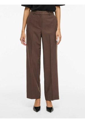 Femme portant un pantalon marron taille haute à jambes larges avec plis sur le devant et des talons noirs pointus, debout devant un fond uni.