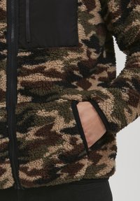 Fleece jacket with a camouflage pattern in green, brown, and black. It features a black zip pocket and soft, textured fabric.