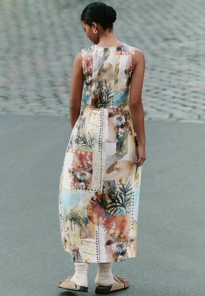 Sieviete ar tumšiem matiem, sakārtotiem bulciņā, valkā bezpiedurkņu midi kleitu ar ziedu un abstraktu rakstu, bēšas zeķes un brūnas slidens kurpes, stāv uz ietves.