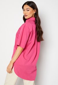 Camicia rosa a maniche corte con colletto, realizzata in tessuto leggero. Presenta un orlo curvo e una vestibilità rilassata, posizionata su uno sfondo semplice.