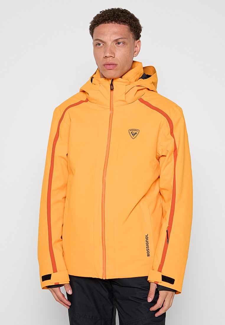Żółta wodoodporna kurtka z pomarańczowymi akcentami, z kapturem, z zamkiem błyskawicznym oraz logo na piersi. Gładka tekstura i sportowy krój.
