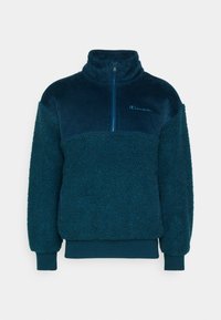 Fleecepullover i mörk turkos med dragkedja i halsen, med ribbade och plyschiga texturer. Inkluderar en liten logotyp på bröstet.