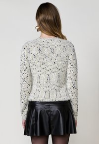 Weißer Pullover mit strukturiertem Muster und goldenen sowie schwarzen Punkten, kombiniert mit einem schwarzen Lederrock. Langärmlig und figurbetont.