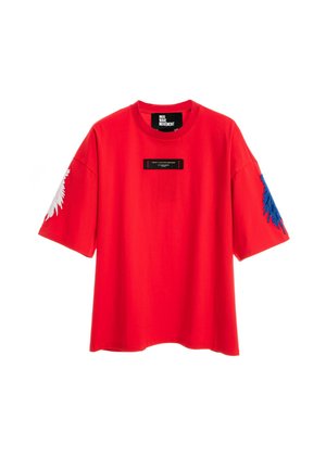 Joven de pie, con camiseta roja brillante de gran tamaño y pantalones cortos a juego con detalle de cadena, calcetines blancos y zapatos beige texturizados sin cordones.