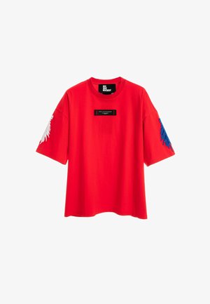 Camiseta roja de manga corta sobredimensionada con etiqueta negra en el pecho y diseños de plumas blancas y azules en las mangas izquierda y derecha.