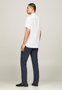 Camicia di lino bianca a maniche corte con yoke posteriore, abbinata a pantaloni blu navy e scarpe nere senza lacci, vista di spalle.