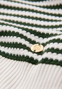 Textil de punto a rayas en colores verde y blanco, con un detalle de emblema circular dorado y una textura acanalada a lo largo del borde inferior.