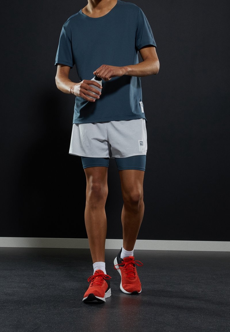 Pánský sportovní outfit se skládá z tmavě modrého trička s krátkým rukávem, světle šedých šortek a červených běžeckých bot se černými a bílými akcenty.