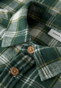 Grønn og hvit rutete flanellskjorte med gule detaljer, med myk tekstur, brettet krage og en lysebrun knapp. Etikett synlig.