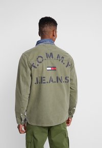 Oliivinvihreä pitkähihaine takki, jossa on farkkukaarroke. Takissa on suuret laivastonsiniset tekstit "T.O.M.M.Y. J.E.A.N.S." ja kolmivärinen lippuembleemi.