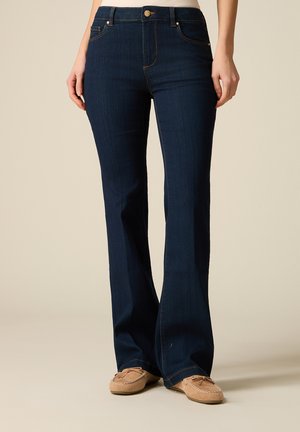 Kobieta ubrana w ciemnoniebieskie dżinsy z rozszerzanymi nogawkami i beżowe mokasyny, stojąca na tle jasnego, gładkiego tła.