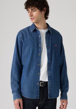 Młody mężczyzna ubrany w niebieską denimową koszulę Levi's na białej koszulce, w połączeniu z czarnymi spodniami i czarnym paskiem, patrzący w bok.