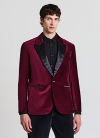 Blazer in velluto bordeaux con rever in satin nero, due tasche frontali e chiusura con un solo bottone. Indossato sopra una camicia nera con bottoni.