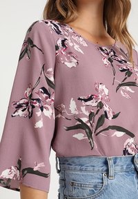 Lila blus med blommönster i rosa, vitt och grönt. Har avslappnad passform och 3/4-långa ärmar, tillverkad av lättviktig tyg.