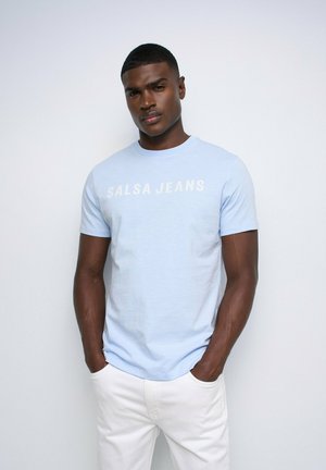 Joven con el pelo corto que lleva una camiseta "SALSA JEANS" azul claro y pantalones blancos, de pie con las manos en los bolsillos frente a un fondo liso.