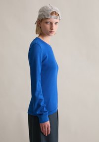 Blaues, langärmliges Rippenoberteil mit Rundhalsausschnitt. Kombiniert mit dunkler Hose und kariertem Cap, zeigt es ein figurnahes Design und eine glatte Textur.