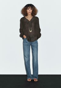 Chemise longue à manches longues en lin marron, à coupe ample, associée à un jean évasé bleu clair. La tenue comprend un long collier avec un pendentif.