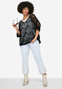 Schwarzer, offener Strickponcho mit Chevron-Muster, über einem weißen Oberteil getragen, kombiniert mit hellblauen Jeans mit runden Ausschnitten und hochgeschlagenen Säumen. Silbern ankle boots.