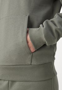 Felpa grigio-verde con tasca a marsupio, polsini e orlo a costine. Tessuto morbido e texturizzato con una vestibilità rilassata.