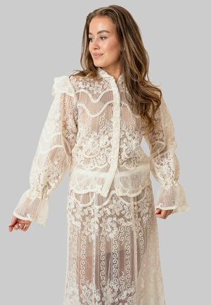 Kobieta o długich brązowych włosach, ubrana w przezroczystą białą koronkową bluzkę i dopasowaną spódnicę, patrząca na bok na tle jednolitego szarego tła.