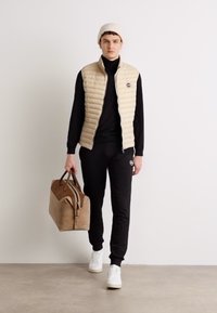 Beige puffertáska fekete garbón, fekete jogger nadrágon és fehér sportcipőn. Könnyűbarna sporttáska van nála. Minimalista design, sima textúrák.