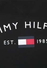 Camisa de algodón negra con el texto blanco "TOMMY HILFIGER," la fecha de establecimiento "EST. 1985," y un logo colorido que se asemeja a una bandera.