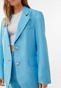 Světle modrý blazer vyrobený z hladkého materiálu. Má klopový límec, dva knoflíky a náprsní kapsu. Volný střih s mírně oversized rukávy.