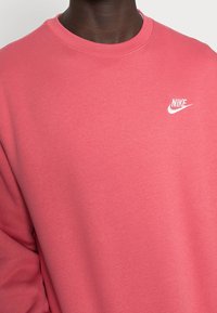 Rosa Nike-hoodie med rund hals, långa ärmar och en liten vit Nike-logotyp på bröstet, tillverkad av mjukt bomullstyg.