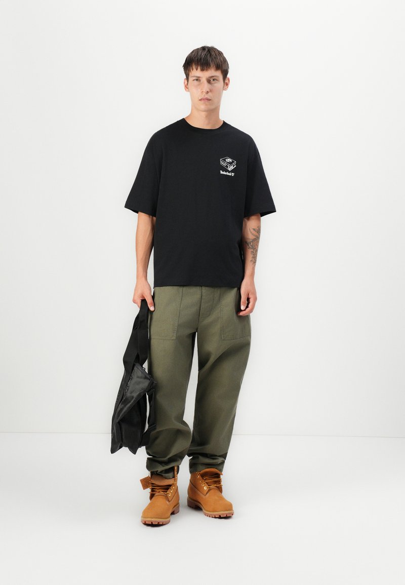 T-shirt nera in cotone con un piccolo logo bianco, pantaloni verde oliva con tasche laterali, stivali marroni e una borsa nera.