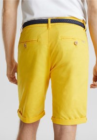 Homme portant un short jaune mi-genou avec des poches arrière boutonnées et une ceinture tissée noire, debout devant un fond uni.
