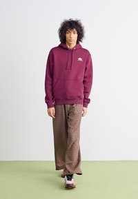 Sweatshirt burdeos con un bolsillo frontal y cordones, combinado con pantalones de cuadros marrones. El modelo lleva zapatillas negras y naranjas. Suelo verde.