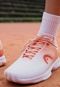 Weiße und hellpfirsichfarbene Tennisschuhe mit strukturierten Oberflächen, einem runden Logo und kontrastierenden Schnürsenkeln, kombiniert mit einer gerippten weißen Socke.