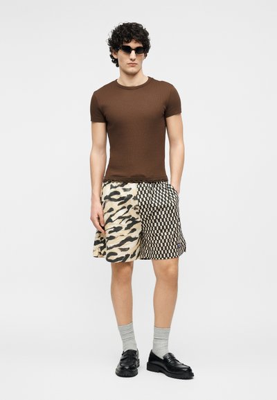 Άνδρας που φοράει καφέ εφαρμοστή μπλούζα, μαύρο-άσπρο σορτς με σχέδιο, γκρι κάλτσες, μαύρα μοκασίνια και μαύρα γυαλιά ηλίου, σε απλό φωτεινό φόντο.