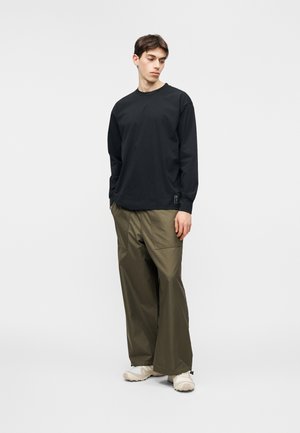 Joven con camisa negra de manga larga, pantalones verdes oliva sueltos y zapatillas beige, mirando hacia abajo con las manos en los bolsillos.