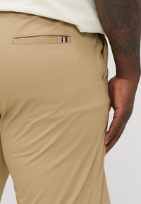 Beige trousers made of smooth fabric, featuring a back pocket with a striped detail in red, white, and blue. The fit appears tailored.