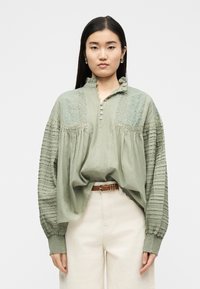 Γυναίκα που φοράει χαλαρό, πράσινο μπλουζάκι φασκόμηλου με δαντέλα και πιετέ λεπτομέρειες, μπροστινό κούμπωμα με κουμπιά, συνδυασμένο με ψηλόμεσο κρεμ παντελόνι και καφέ ζώνη.