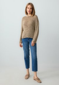 Beige gestrickter Pullover mit lockerer Passform und weiten Ärmeln, kombiniert mit blauen Jeans im geraden Schnitt und hellbraunen Slippers.