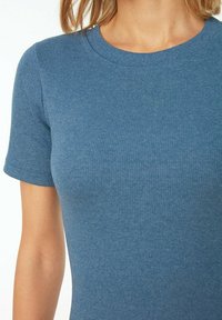 Kvinna klädd i en figurnära, blå ribbad kortärmad tröja med rund halsringning, visad från axlarna till mitten av bålen.