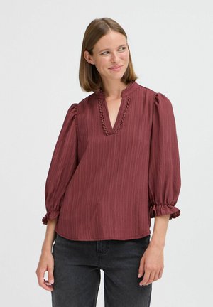 Bordeauxfarbige Bluse mit gepufften Ärmeln, strukturiertem Stoff und einem V-Ausschnitt, der mit einer gewellten Borte verziert ist. Kombiniert mit dunklen Jeans.