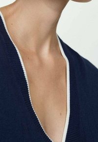 Vêtement tricoté marine avec un décolleté en V profond et des bordures blanches contrastantes. Présente une texture douce et des détails en côtes le long du col.