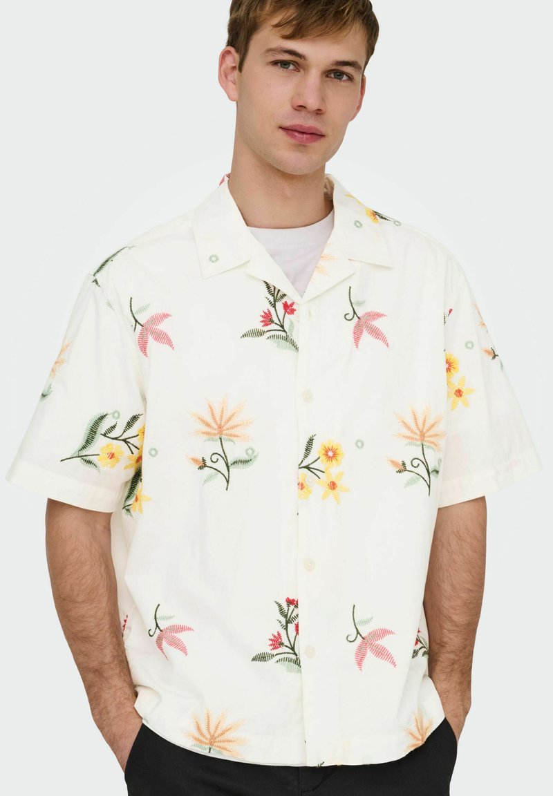 Mladý muž v bielej krátkorukávovej košeli s farebnými kvetinovými vzormi, stojaci s rukami v vreckách na jednoliatom pozadí.