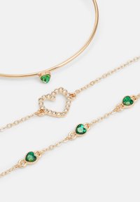 Set di gioielli in chain color oro con design a forma di cuore, impreziosito da strass verdi e trasparenti per un ulteriore dettaglio.