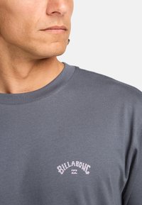 Grå bomulds t-shirt med rund hals, der har et lille hvidt "Billabong" logo på brystet. Glat tekstur, afslappet design.