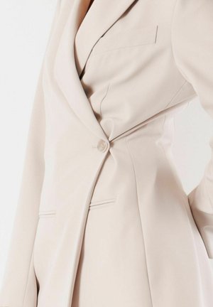 Primo piano di un blazer beige su misura abbottonato in vita, che mostra il risvolto, i dettagli delle tasche e la texture liscia del tessuto.