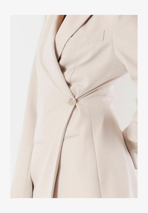 Primo piano di un blazer beige su misura abbottonato in vita, che mostra il risvolto, i dettagli delle tasche e la texture liscia del tessuto.