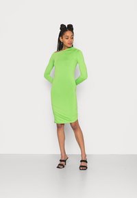 Vestido bodycon verde com mangas longas, apresentando um decote à mostra nos ombros e textura franzida na cintura, combinado com saltos pretos.