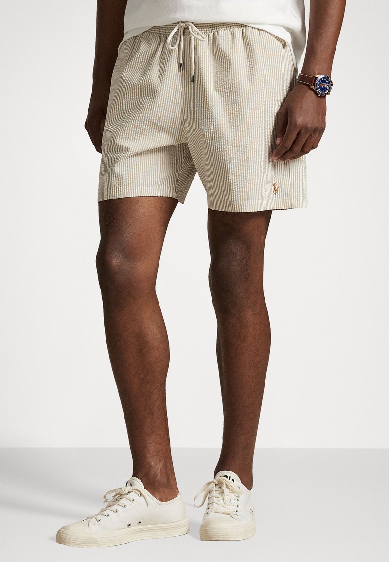 Beige Strickshorts mit vertikalen Streifen, elastischer Taille mit Kordelzug und kleinem gesticktem Logo. Kombiniert mit weißen Leinenschuhen.