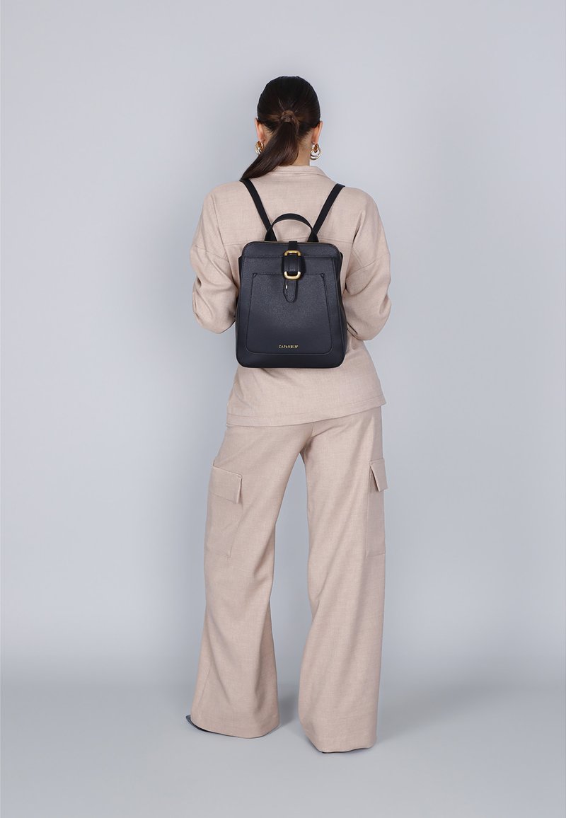 Sac à dos en cuir noir de forme rectangulaire, avec un détail de boucle à l'avant et des bretelles ajustables, porté par une personne de dos.