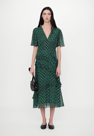 Zelené vzorované šaty s krátkými volánovými rukávy, výstřihem do V a vrstvenými volánky; nošené s malou černou kabelkou a balerínami.
